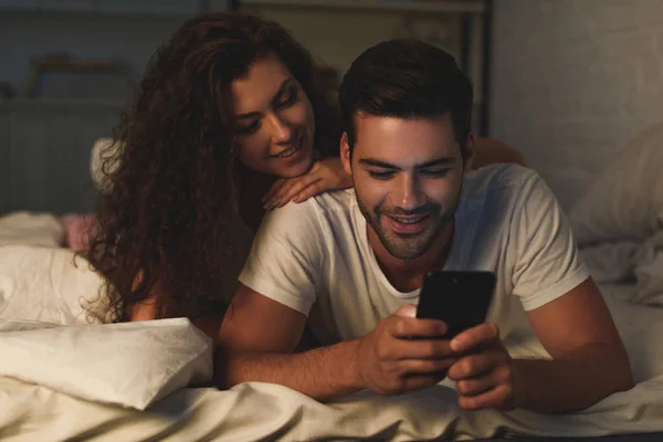 Feliz jovem casal deitado na cama e usando smartphone juntos — Fotografia de Stock