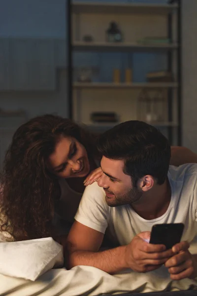 Happy young couple smiling each other while lying on bed and using smartphone — Stock Photo