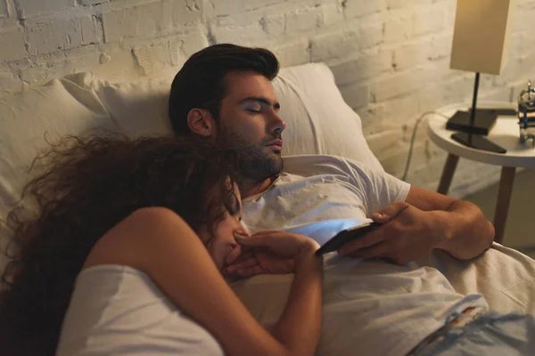 Vue grand angle de la jeune femme qui dort tout en petit ami en utilisant un smartphone au lit — Photo de stock