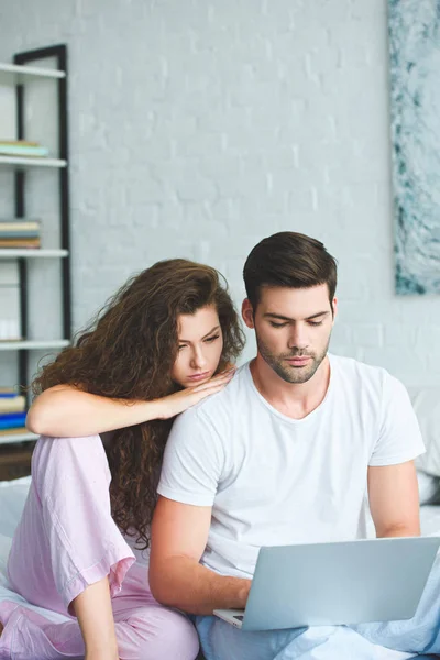 Ernsthafte junge Paar im Pyjama mit Laptop auf dem Bett — Stockfoto