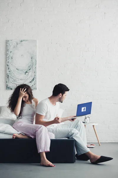 Upset young woman sitting on bed while angry man using laptop with facebook website on screen — Stock Photo