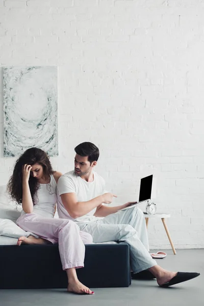 Mujer joven molesta sentada en la cama mientras que el hombre señala con el dedo a la computadora portátil con pantalla en blanco - foto de stock