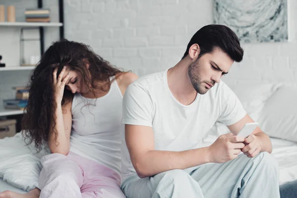 Jeune homme utilisant smartphone tandis que la femme bouleversée assis derrière sur le lit, concept de difficultés relationnelles — Photo de stock