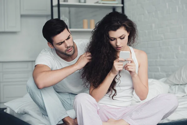 Chateado jovem olhando para a namorada usando smartphone na cama, conceito de dificuldades de relacionamento — Fotografia de Stock