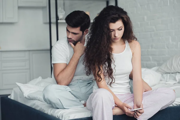 Young woman using smartphone while upset man sitting behind on bed, relationship difficulties concept — Stock Photo