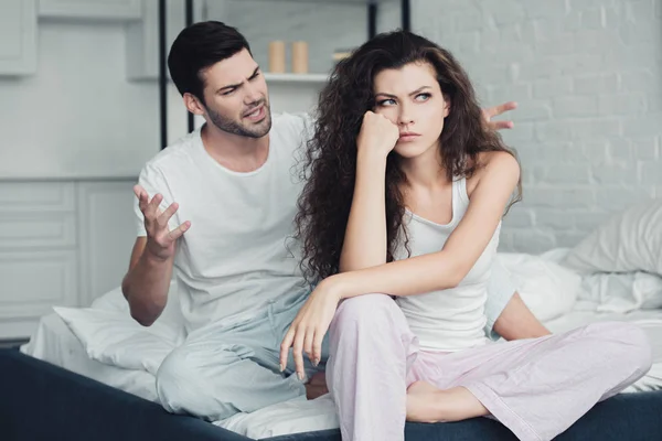 Jeune couple en pyjama assis sur le lit et querelles, concept de difficultés relationnelles — Photo de stock