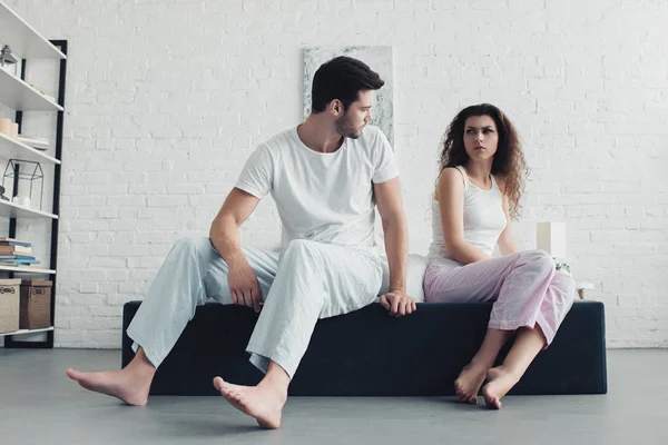 Bouleversé jeune couple en pyjama assis sur le lit et se regardant, concept de difficultés relationnelles — Photo de stock