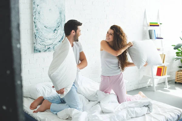 Vista lateral de feliz pareja joven divirtiéndose y luchando con almohadas en la cama - foto de stock
