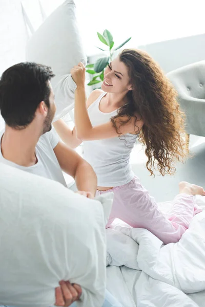 Feliz joven pareja divirtiéndose y luchando con almohadas en la cama - foto de stock