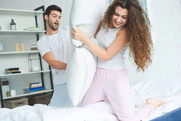 Feliz joven pareja en pijama divertirse y luchar con almohadas en la cama - foto de stock