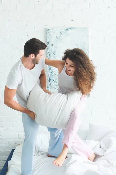 Happy young couple in pajamas fighting with pillow on bed — Stock Photo