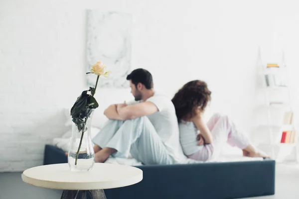 Rose flower in vase and upset young couple sitting back to back on bed behind — Stock Photo
