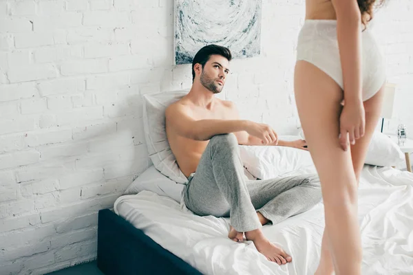 Обрезанный снимок красивого мужчины, смотрящего на чувственную девушку в нижнем белье, стоящую на кровати — Stock Photo