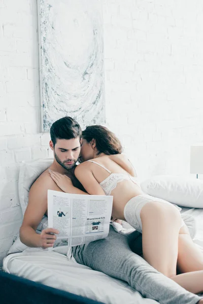 Sexy girl in lingerie kissing boyfriend reading newspaper in bed — Stock Photo