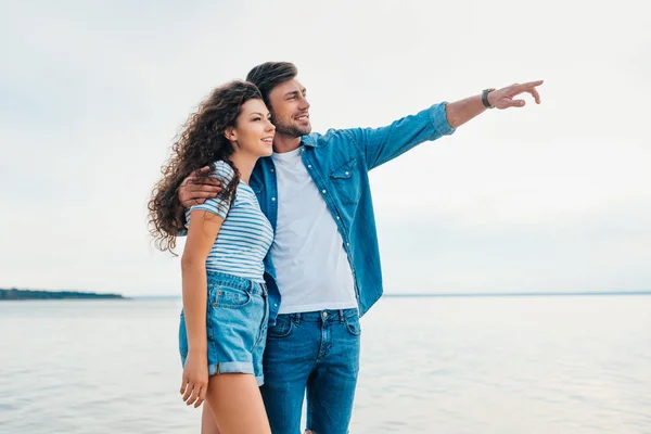 Couple heureux étreignant sur la plage et pointant vers la mer — Photo de stock