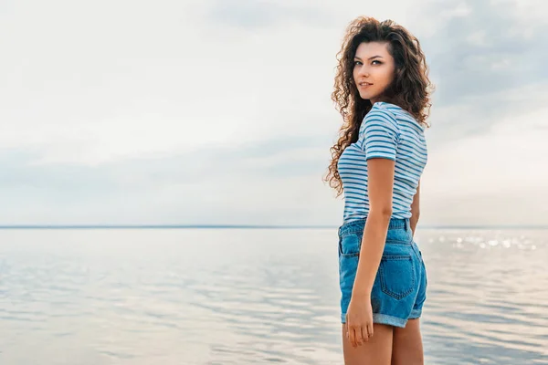 Приваблива кучерява дівчина в повсякденному одязі позує біля морів — стокове фото