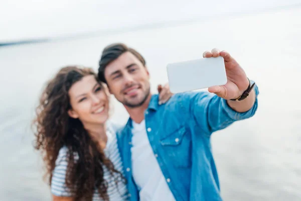 Enfoque selectivo de la pareja sonriente tomando selfie en el teléfono inteligente cerca del mar - foto de stock