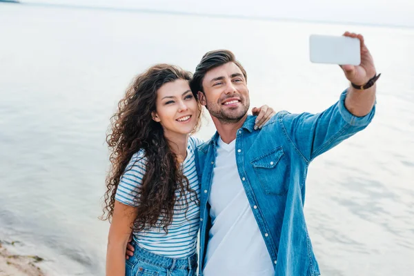 Joven sonriente pareja tomando selfie en smartphone cerca del mar - foto de stock