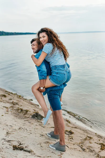 Cheerful young couple piggybacking on beach near sea — Stock Photo