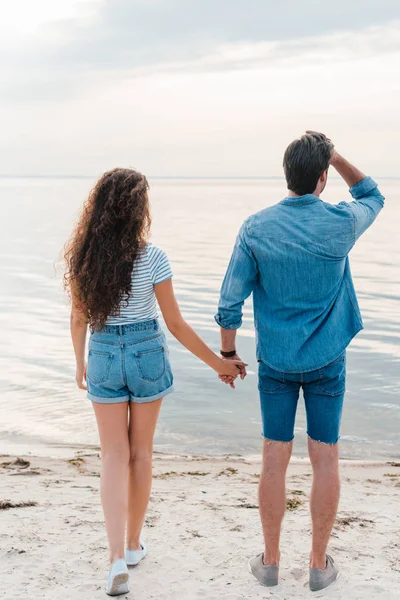 Rückansicht eines jungen Paares, das sich an den Händen hält und aufs Meer blickt — Stockfoto