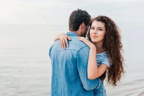 Joven sonriente pareja abrazándose en la playa cerca del mar - foto de stock