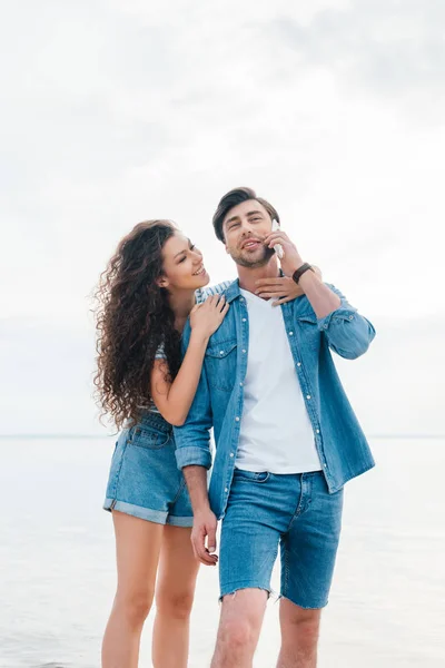 Young girlfriend hugging her boyfriend while he talking on smartphone near sea — Stock Photo