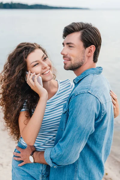 Hombre abrazando a su novia sonriente mientras ella habla en el teléfono inteligente cerca del mar — Stock Photo