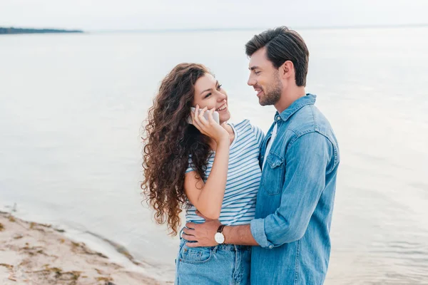 Giovane uomo che abbraccia la sua ragazza mentre lei parla su smartphone vicino al mare — Foto stock