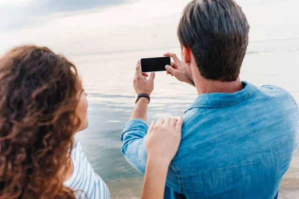 Vista posterior de la pareja tomando fotos del mar en el teléfono inteligente - foto de stock
