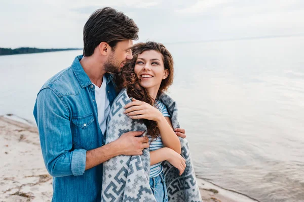 Boyfriend embracing smiling girlfriend wrapped in blanket near sea — Stock Photo