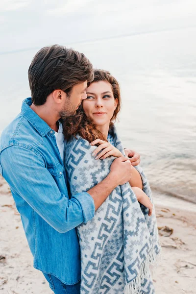 Jeune homme câlin attrayant copine enveloppé dans une couverture sur la plage — Photo de stock