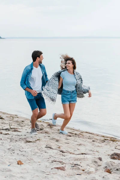 Молодая улыбающаяся пара, держащаяся за руки и бегущая по берегу моря — стоковое фото