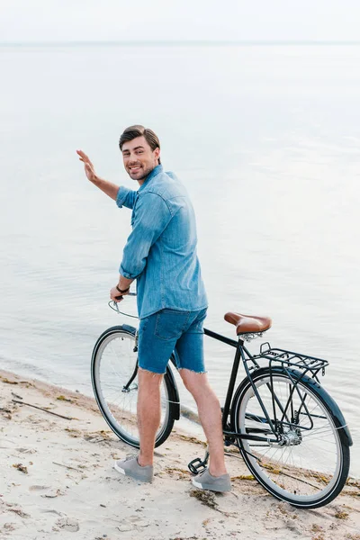 Felice uomo che saluta e cammina con la bicicletta sulla spiaggia di sabbia — Foto stock