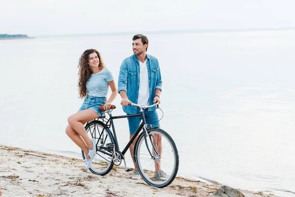 Smiling couple sitting on bike on beach near sea — Stock Photo