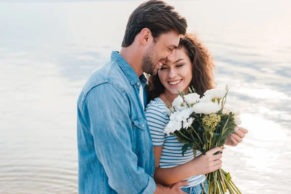 Smiling couple embracing and holding bouquet near the sea — Stock Photo