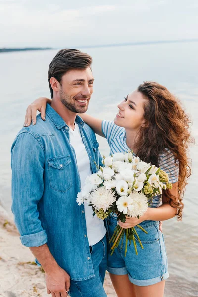 Schönes junges Paar umarmt und hält Blumenstrauß am Meer — Stockfoto