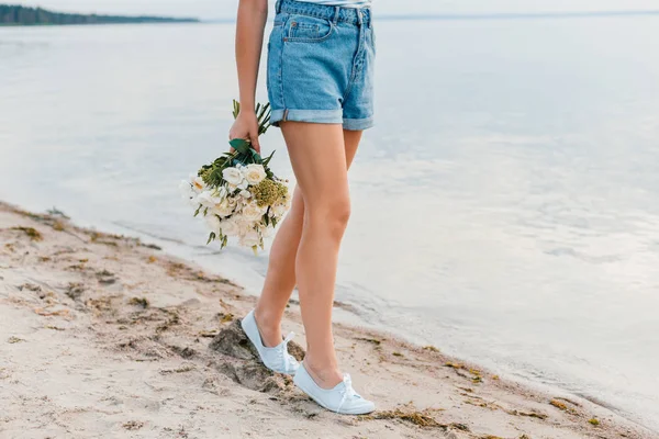 Vista de sección baja de chica con ramo de caminar en la playa cerca del mar - foto de stock