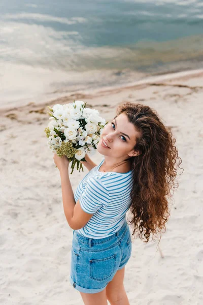 Belle femme bouclée souriante avec bouquet posant sur le bord de la mer — Photo de stock
