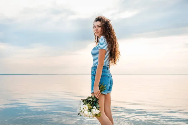 Attraente giovane donna con mazzo di fiori in posa vicino al mare — Foto stock
