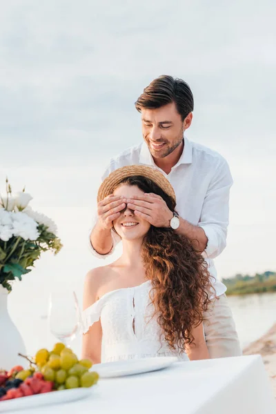 Sonriente hombre cerrando los ojos y haciendo sorpresa para la novia, cita romántica en la orilla del mar - foto de stock