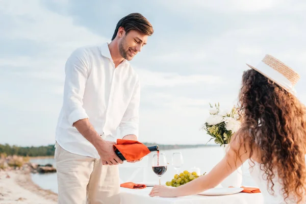 Happy man pouring red wine into glass for woman on romantic date on beach — Stock Photo