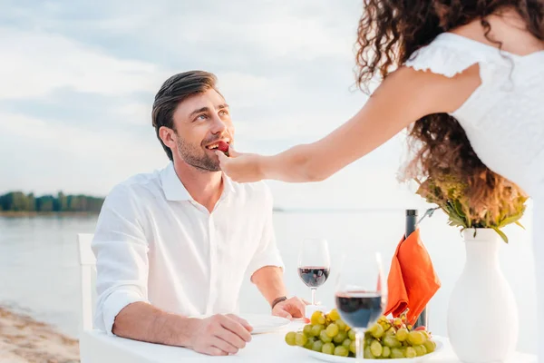 Chica alimentación feliz novio con fresa durante romántica fecha cerca del mar - foto de stock