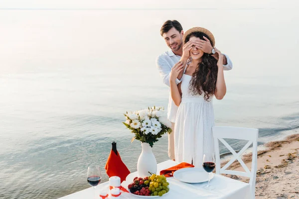 Hombre feliz cerrando los ojos y haciendo sorpresa para la novia, cita romántica cerca del mar - foto de stock