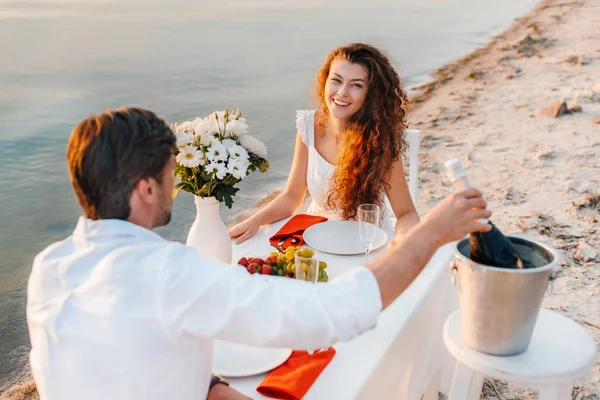 Мужчина берет бутылку шампанского для романтического свидания на пляже с улыбающейся девушкой — стоковое фото