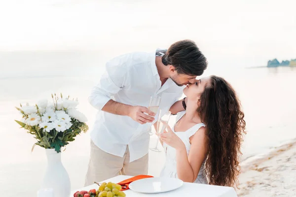 Feliz pareja besando y sosteniendo copas de champán durante una cita romántica en la orilla del mar - foto de stock