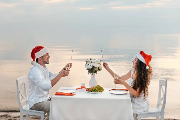 Feliz pareja en sombreros de santa celebrando la Navidad con copas de champán y bengalas en la playa - foto de stock