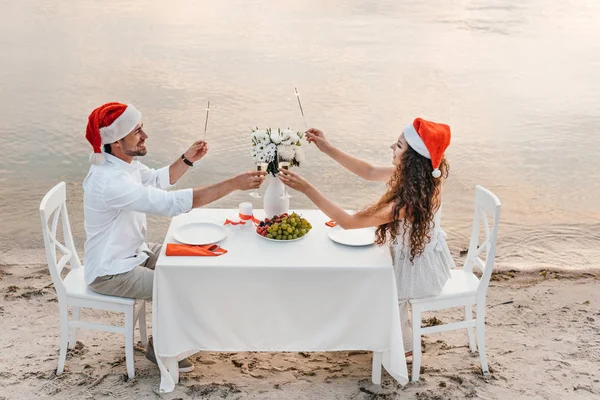 Pareja joven en sombreros de santa celebración de bengalas y tostadas con copas de champán mientras se celebra la Navidad en la playa - foto de stock