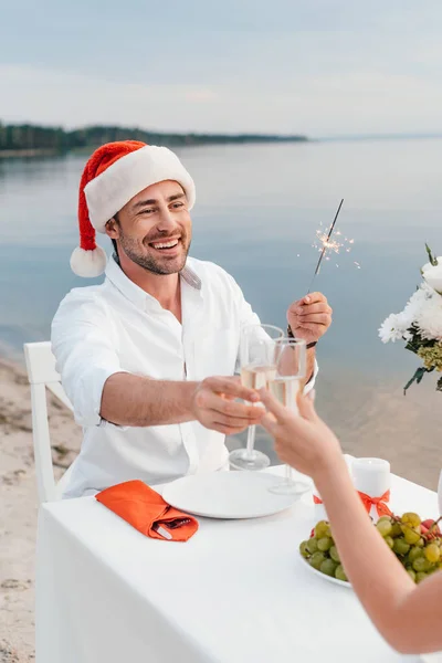 Feliz pareja celebrando la Navidad y tintineo copas de champán en la playa - foto de stock