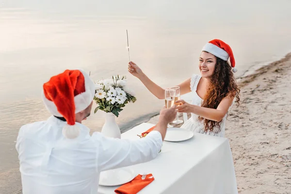 Pareja joven en sombreros de santa celebración de Navidad y tintineo con copas de champán en la playa - foto de stock