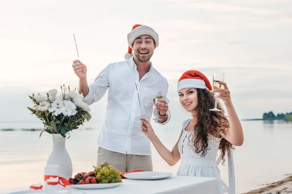 Pareja de sombreros de santa celebrando el año nuevo con copas de champán y bengalas en la playa - foto de stock